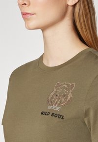 Camiseta verde olivo de mangas cortas, con un diseño de león adornado con pedrería y el texto "ALMA SALVAJE" debajo de la gráfica.