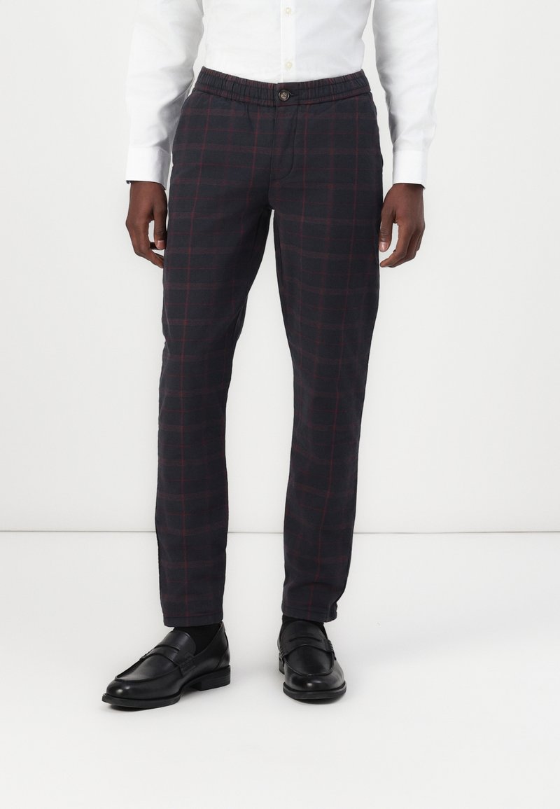 Pantalones de cuadros gris oscuro, con una cintura relajada, detalles rojos en el patrón de cuadros y un diseño ajustado. Combinados con mocasines negros.