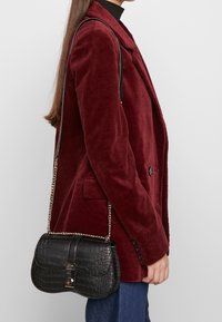 Czarna torebka crossbody z ekoskóry o teksturowanym wzorze krokodyla, z metalowymi elementami i łańcuszkowym paskiem, trzymana przez osobę ubraną w burgundowy żakiet.