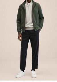 Zielona kurtka bomberka na białym swetrze z zamkiem, w zestawieniu z ciemnymi spodniami i białymi trampkami. Prosty design w neutralnych odcieniach.