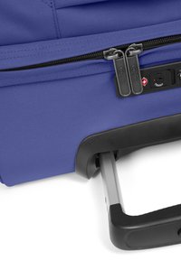 Valigia in nylon viola con scomparto con zip, manico telescopico e dettagli neri. Dotata di una serratura combinata e superficie testurizzata.