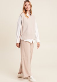 Beige gebreide vest over een witte blouse, gecombineerd met lichtroze gestreepte wijde broek en witte sneakers. Eenvoudig en modern ontwerp.