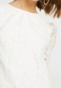 Hvid bluse i blonder med rund halsudskæring og korte ærmer. Stoffet har et blomstermønster og en tekstureret overflade.