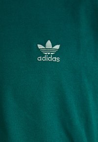 Grøn t-shirt lavet af blødt stof med et lille hvidt broderet Adidas-logo centreret nær nakken, med en ribbet tekstur.