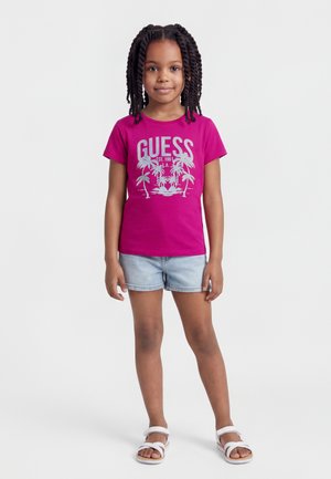 Fille aux cheveux tressés portant un t-shirt GUESS rose vif, un short en jean bleu clair et des sandales blanches, se tenant devant un fond uni.