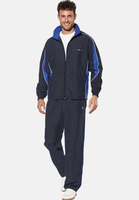 Marineblauer Sportanzug mit blauen Akzenten, Reißverschlussjacke, elastischen Bündchen und passenden Hosen. Getragen mit weißen Turnschuhen, glatte Stofftextur.