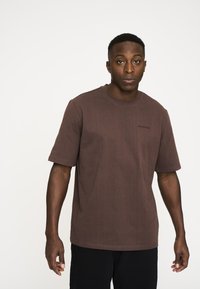 Brun bomulds T-shirt med rund hals og korte �ærmer. Har et diskret mærkelogo på den øverste venstre side. Enkel design.