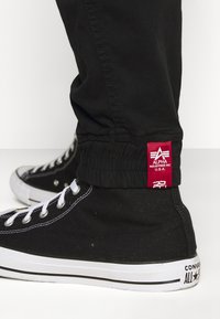 Svart joggebukser med elastisk mansjett og et rødt Alpha Industries-merke. Kombinert med svarte høyhælte Converse-sneakers med hvite snøring og detaljer.