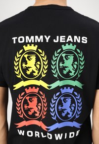 Fekete póló "TOMMY JEANS" felirattal. Négy színes címer (sárga, zöld, piros, kék) oroszlánokkal és babérlevelekkel, valamint egy transzparenssel.