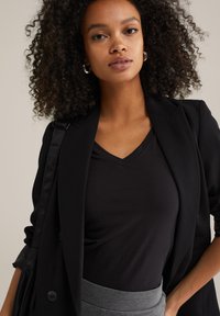 Femeie cu păr creț, purtând cercei din argint în formă de cerc, sacou negru, top negru cu decolteu în V, pantaloni gri și purtând o geantă de umăr neagră.