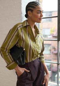Olijfgroen gestreept blouse met opgerolde mouwen, gecombineerd met donkere broek; holding een zwarte handtas. Helder, natuurlijk licht van een raam.