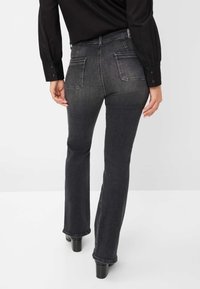 Jeans noirs évasés avec un lavage dégradé, présentant des poches arrière et une texture subtile. Le mannequin porte des talons noirs et une chemise noire retroussée.