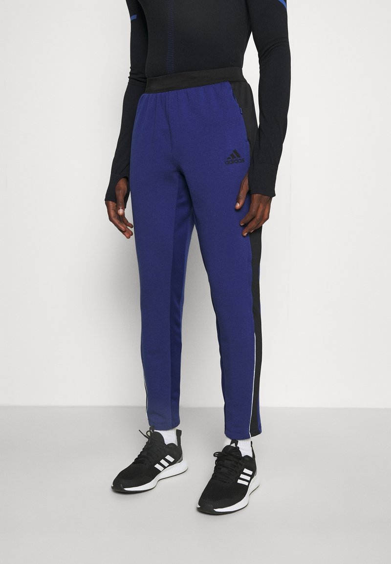 Pantalon de sport bleu avec une taille noire, des bandes latérales et des poches zippées. Tissu texturé et logo Adidas subtil sur la cuisse gauche.