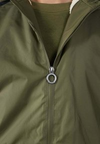 Olijfgroene jas met een gladde, glanzende stof en een fleece voering aan de binnenkant. Voorzien van een rits aan de voorkant met een ronde metalen trekker en een hoge kraag.