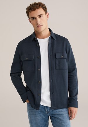 Marineblaues Hemd mit Knopfleiste und zwei Brusttaschen, aus strukturiertem Material, kombiniert mit einem weißen T-Shirt und getragen mit blauen Jeans.