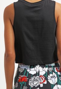 Person som bär en ärmlös svart crop top och byxor med blommönster i vitt, rött och grönt på en mörk bakgrund.