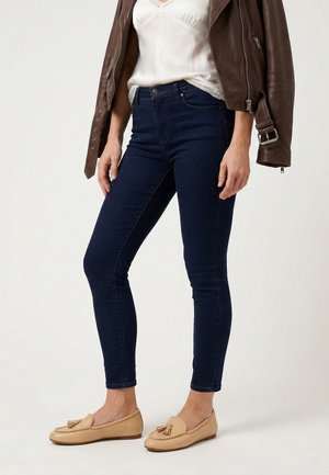 Vrouw draagt donkerblauwe slim-fit spijkerbroek, beige loafers met kwastjes, witte top met kanten rand en een bruine leren jas losjes over de schouders gedragen.