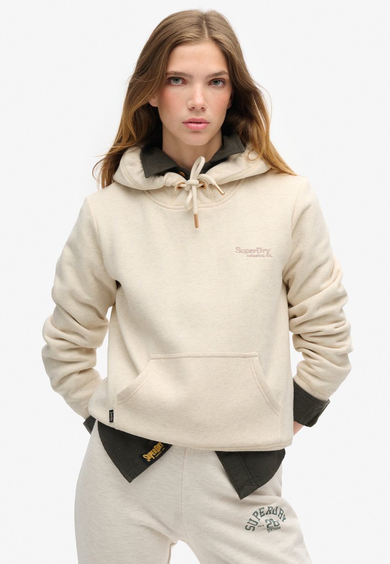 Felpa con cappuccio crema, dotata di tasca frontale, chiusura con cordoncino e logo ricamato. Abbinata a pantaloni tuta beige con logo verde.