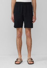 Shorts noirs texturés avec une taille élastique et un cordon de serrage. Portés avec des sandales marron à double boucle.