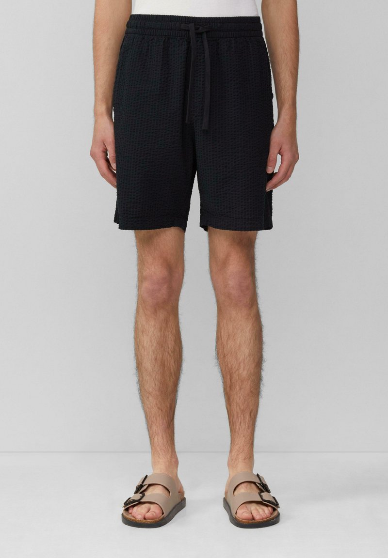 Shorts noirs texturés avec une taille élastique et un cordon de serrage. Portés avec des sandales marron à double boucle.