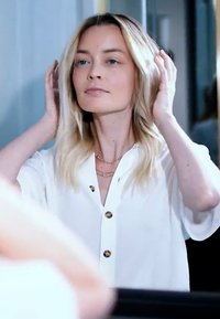 Blonde vrouw in een witte blouse die haar haar netjes maakt terwijl ze naar haar spiegelbeeld kijkt binnenshuis.