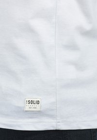Camiseta de algodón blanca con una pequeña etiqueta negra en el dobladillo que muestra el texto "!SOLID DENMARK EST.1999." Textura suave, ajuste estándar.