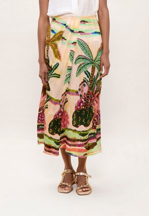 Mujer con una falda midi colorida con estampado de palmeras tropicales y sandalias abiertas con adorno dorado, de pie frente a un fondo liso.