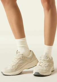 Beige sportsko i tekstureret mesh og ruskindsdele, med flade snørebånd og en hvid gummisål. Båret med hvide ribbede sokker.
