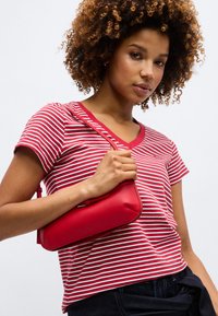 Γυναίκα με σγουρά μαλλιά που φοράει κόκκινο και άσπρο ριγέ μπλουζάκι με V-λαιμόκοψη, κρατώντας μια μικρή κόκκινη δερμάτινη τσάντα ώμου, μπροστά σε απλό φόντο.