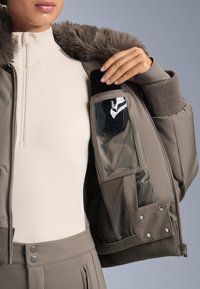 Barna bombázó dzseki műszőrme gallérral, bordázott mandzsettákkal és belső zsebbel, amely átlátszó panellel rendelkezik a láthatóság érdekében.