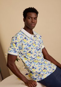 Korte mouwen polo-shirt met een citroen- en bladerenpatroon in geel, blauw en groen op een witte achtergrond; afgewerkt met een witte kraag.
