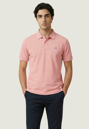 Hombre con camiseta polo de manga corta rosa con pequeño logo y pantalones azul oscuro, de pie con las manos en los bolsillos frente a un fondo liso.