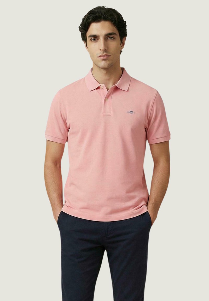 Uomo che indossa una polo rosa a maniche corte con un piccolo logo e pantaloni blu scuro, in piedi con le mani in tasca davanti a uno sfondo semplice.