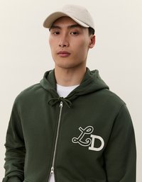 Zelená mikina na zips s prednou kapsou, kapucňou so šnúrkou a bielym logom "LD". Šedá čiapka s výšivkou. Jednoduché pozadie.