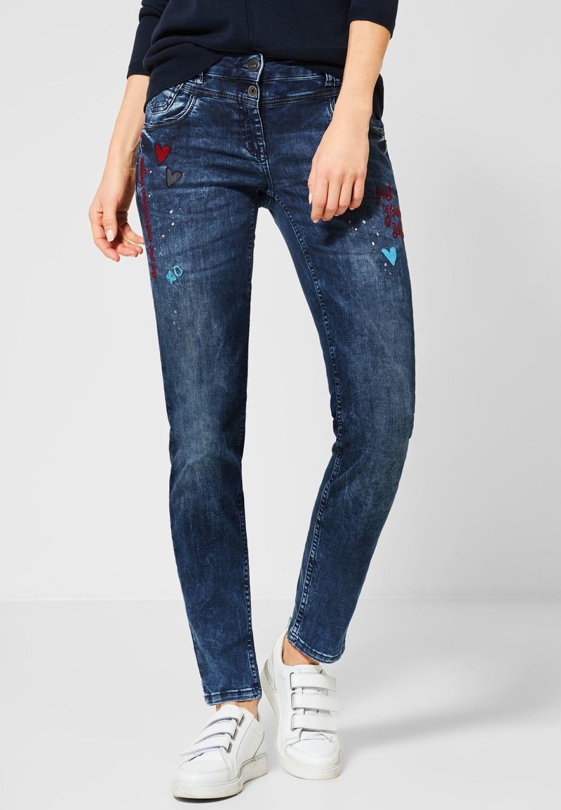 Femme portant un jean skinny bleu foncé avec broderie cœur et "XO", associé à des baskets blanches à scratch sur un fond uni.