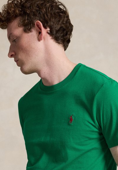 Ung mand med krøllet hår iført en grøn crewneck T-shirt med en lille rød broderet logo på brystet, kigger til venstre.