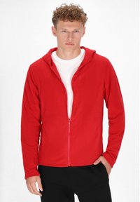 Felpa rossa con zip, dotata di maniche lunghe e cappuccio, indossata sopra una maglietta bianca. Texture liscia e vestibilità casual.