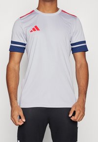 Grå atletisk skjorta med röda axelränder och marinblå ribbade ärmar. Har en Adidas-logotyp på vänster bröst. Slät tygstruktur.