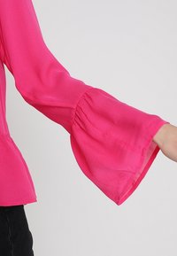Blusa rosa aderente con maniche lunghe svasate e trasparenti e dettaglio arricciato ai polsi. Tessuto liscio con una texture leggera.