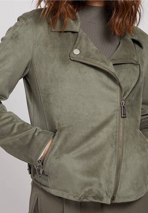 Kobieta w oliwkowo-zielonej zamszowej kurtce motocyklowej z asymetrycznym zamkiem błyskawicznym i rękami w kieszeniach bocznych na zamek, nosząca pod spodem prążkowany sweter.