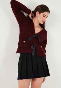 Burgundy knitted cardigan with cable patterns, v-neck, and black ribbon ties at the waist; paired with a black pleated skirt.