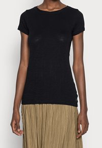 Czarny, dopasowany t-shirt z krótkim rękawem połączony z beżową spódnicą plisowaną, na osobie stojącej na jednolitym tle.