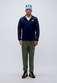 Άνδρας που στέκεται φορώντας ναυτικό μπουφάν, χακί παντελόνια, μοβ σκουφάκι, μπλε ανακλαστικά γυαλιά ηλίου και παπούτσια πεζοπορίας σε λευκό φόντο.