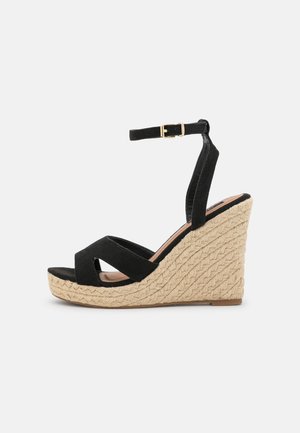 Sort ruskind kile sandal med ankelrem og guldbeslået spænde, med flettet beige plateauhæl.