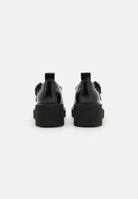 Black patent leather shoes with a chunky black sole, featuring a pull tab and decorative bow detail on the side.
