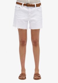 Shorts en denim blanc avec un ourlet effiloché, dotés de passants de ceinture. Associés à une ceinture tressée marron et des sandales à glisser marron sur des pieds nus.