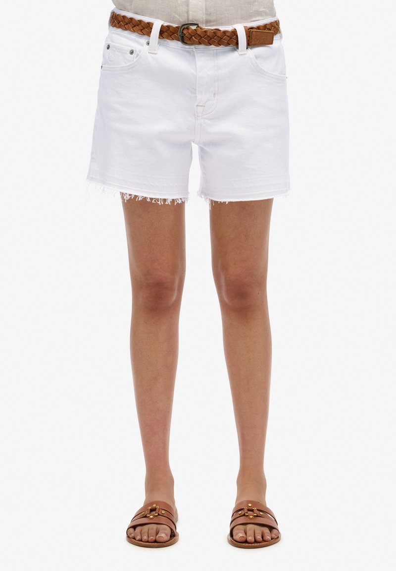 Shorts en denim blanc avec un ourlet effiloché, dotés de passants de ceinture. Associés à une ceinture tressée marron et des sandales à glisser marron sur des pieds nus.