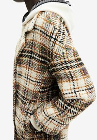Cappotto a quadri in misto lana beige, marrone e nero. Presenta delle macchie multicolori, ampie tasche e un colletto a costine bianco e nero.