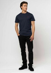 Fiatal férfi sötétkék pólót visel piros logóval, fekete nadrágban és fekete cipőben áll, kezét zsebébe dugva, az oldalra néz.