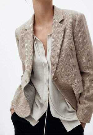 Femme portant un blazer chevrons beige sur une chemise en lin claire boutonnée, les mains dans les poches d'un pantalon noir.